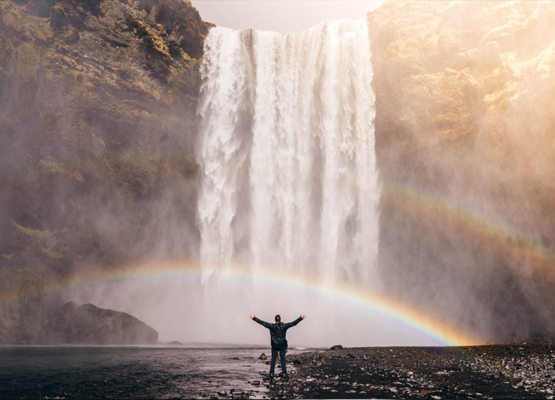 Photo of a person standing in front of a waterfall -created rainbow.